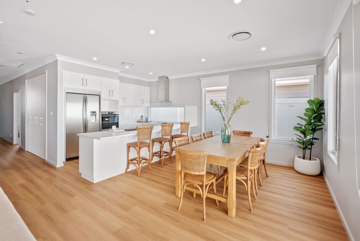 Modern kitchen and dining area with light wood flooring, white cupboards, stainless steel appliances, a wooden dining table for six, and a large plant in the corner. Filled with natural light and open for viewing.