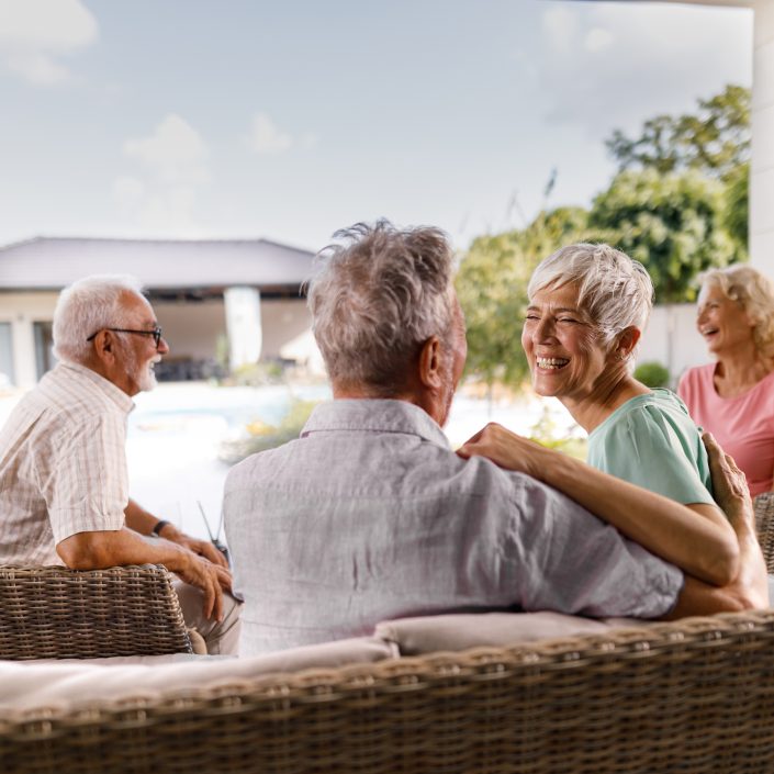 Four older adults sit on wicker sofas outdoors, smiling and laughing together during an Information Session, enjoying a sunny day. A house and greenery in the background create a relaxed and cheerful atmosphere.