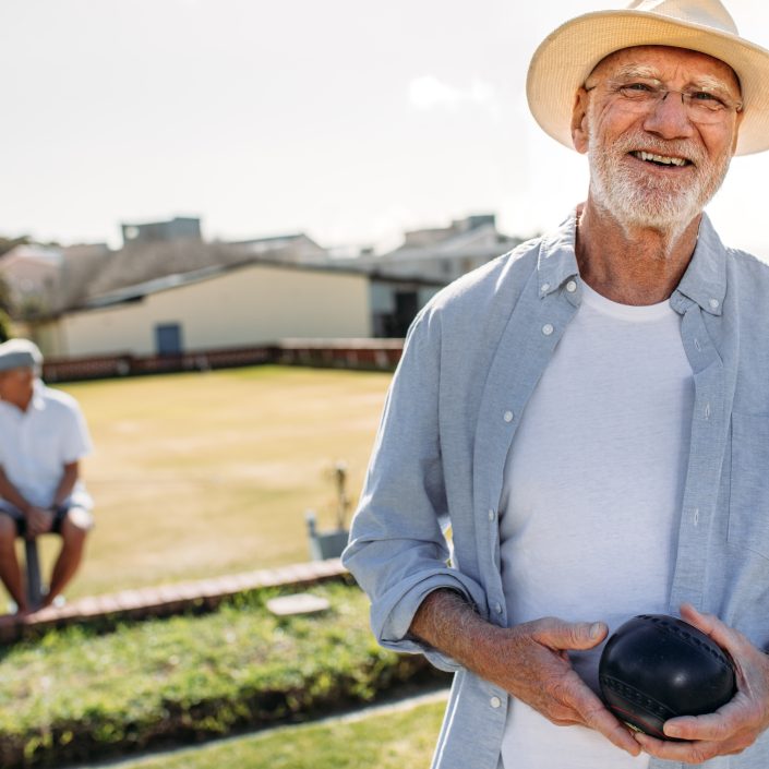 An elderly man in a hat and glasses smiles whilst holding a lawn bowl on a sunny day at an Information Session, with two other people sitting on a bench and a bowling green in the background.