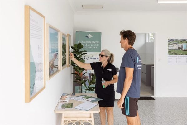 A woman in black attire points to framed posters on a wall and discusses new lifestyle amenities at Northern Rivers Lifestyle Estate with a man in casual clothes inside a bright office. A table with brochures and a banner are visible in the background.