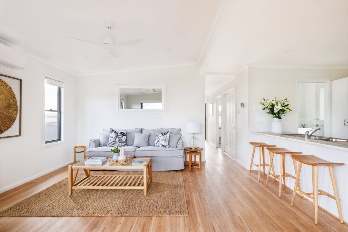 Bright, modern living room maximising your space in a downsized home—light wood floors, grey sofa, patterned cushions, wooden coffee table, jute rug, and barstools at a white kitchen counter. Large wall mirror and potted plant add decor.