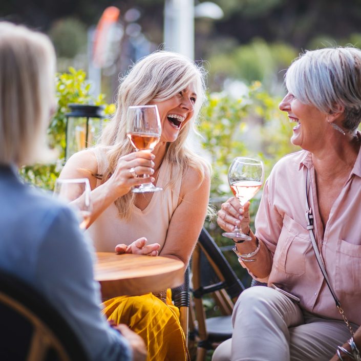 Three women with short blonde and grey hair sit at an outdoor table on a sunny decking, laughing and holding glasses of rosé wine as they enjoy a lively conversation.