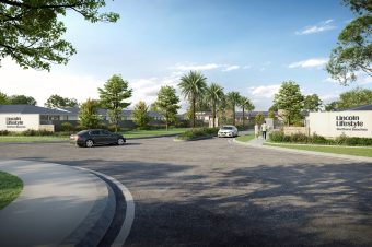A sunny entrance welcomes you to Lincoln Lifestyle Northern Beaches, featuring two cars on a wide road lined with palm trees, lush landscaping, and elegant stone walls displaying the community’s name.