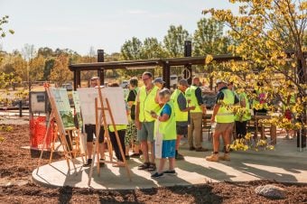 A group of people wearing yellow safety vests gather outdoors around information boards set up on easels, with trees and the Baranduda Lifestyle Estate Clubhouse nearing completion in the background on a sunny day.