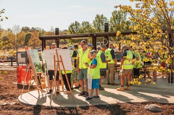 A group of people wearing yellow safety vests gather outdoors around information boards set up on easels, with trees and the Baranduda Lifestyle Estate Clubhouse nearing completion in the background on a sunny day.