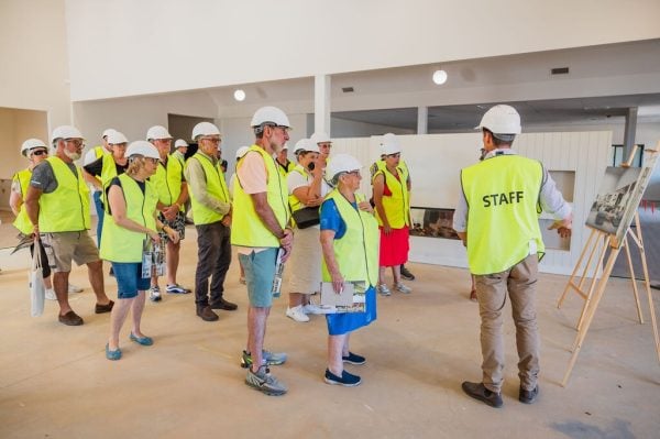 A group of people wearing yellow safety vests and white hard hats listen to a staff member explaining details from an easel during a building tour as the Baranduda Lifestyle Estate Clubhouse nears completion inside the construction site.