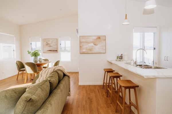 Bright, modern open-plan living space with a green sofa, wooden dining table and chairs, bar stools at the kitchen counter, large windows, and minimalist decor in neutral tones. Saturday Open for Viewing—don’t miss your chance to view!.