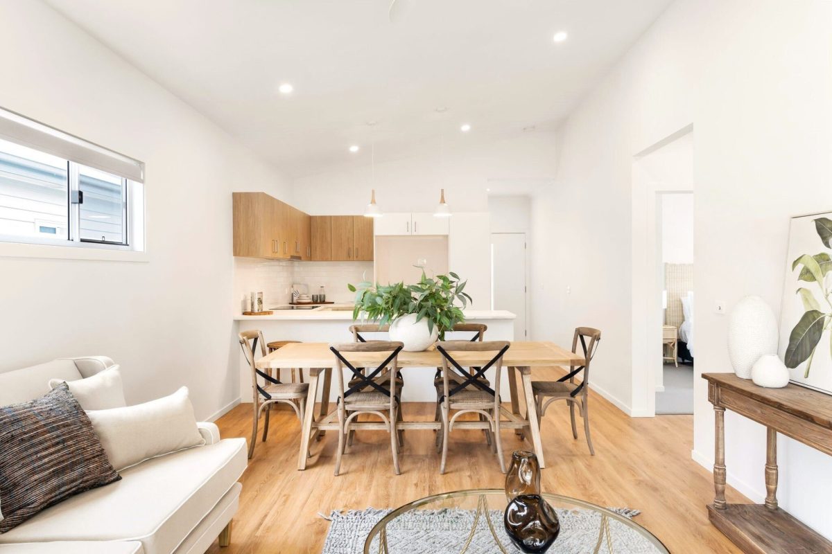 Bright, modern open-plan dining and kitchen area with wooden floors, a wooden table for six, potted plant centrepiece, white cabinetry, and minimalist decor in neutral tones. Saturday Open for Inspection available—don’t miss your chance to view!.