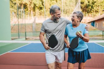 An older man and woman smile and laugh together on a pickleball court, holding paddles and a ball, with an outdoor net and trees in the background—showing how Lincoln Place supports a healthy lifestyle through fun, active living.