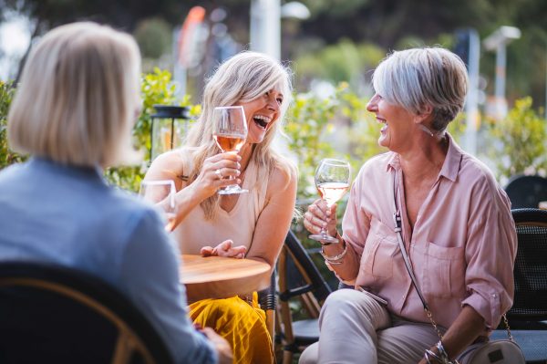 Three women with fair hair sit at an outdoor table, smiling and laughing while toasting with glasses of rosé wine on a cheerful afternoon. The bright, green setting adds to the relaxed atmosphere.