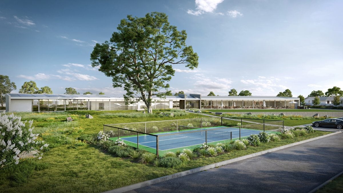 A modern building with large windows sits behind a fenced tennis court and lush green landscaping under a blue sky at Eagle Point, with a large tree and parked cars nearby—perfect for enjoying the Lincoln Lifestyle.