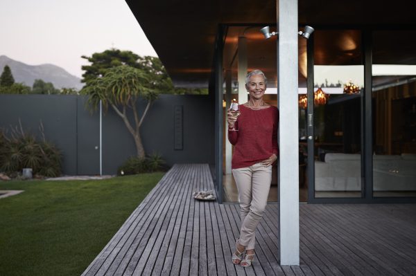 An older woman with short grey hair stands on a wooden decking outside a modern house, smiling and holding a glass of wine. Dressed in a red jumper, she enjoys Priority Access Club benefits amid scenic trees and mountain views in the background.