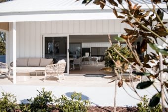A modern patio with wicker furniture and cushions sits before a white building with large sliding glass doors, showcasing inviting interiors. Green shrubs and a small tree complete this relaxing space—ideal for enjoying Hunter Valley over 50s amenities.