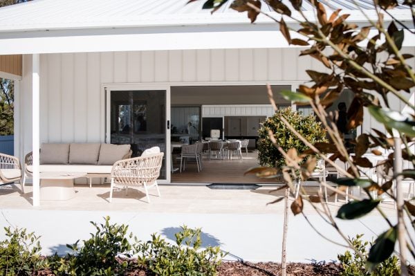 A modern patio with wicker furniture and cushions sits before a white building with large sliding glass doors, showcasing inviting interiors. Green shrubs and a small tree complete this relaxing space—ideal for enjoying Hunter Valley over 50s amenities.