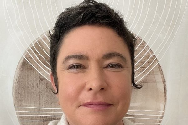 Rebecca Woodleigh, with short dark hair and light skin, smiles gently at the camera, wrapped in a textured blanket with geometric patterns. Abstract circular artwork with white lines is visible on the wall behind her.