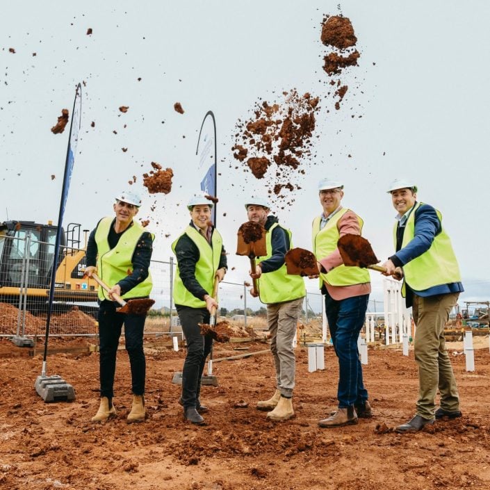Five people wearing hard hats and yellow safety vests smile as they toss shovels of soil at the Griffith community over-50s clubhouse construction site, with soil flying and equipment visible in the background.