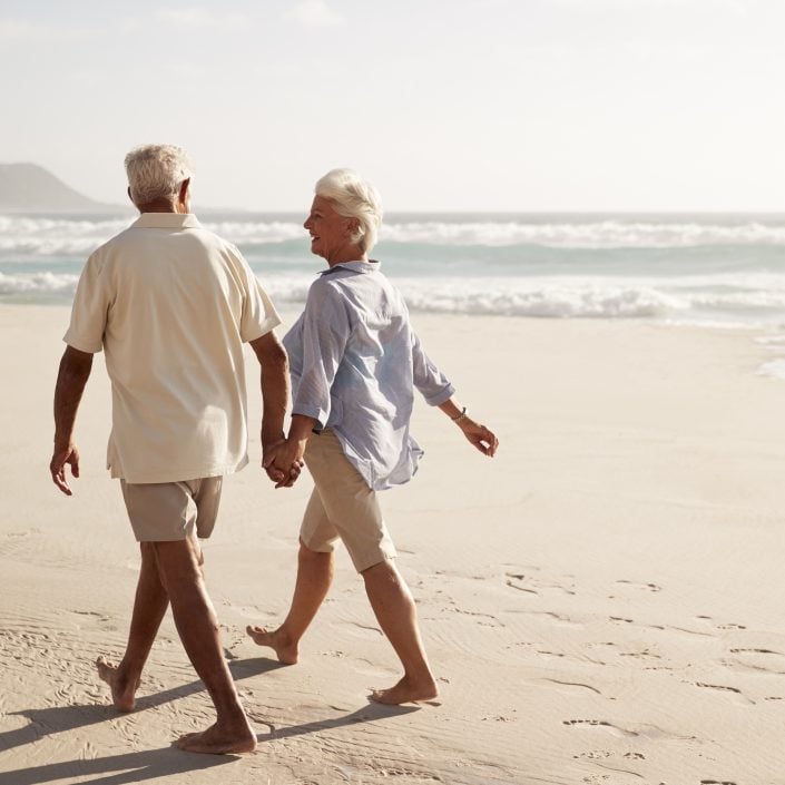 An elderly couple walk hand in hand along a sandy Yeppoon beach by the sea, smiling at each other with waves in the background on a sunny day, enjoying the relaxed Lincoln lifestyle.