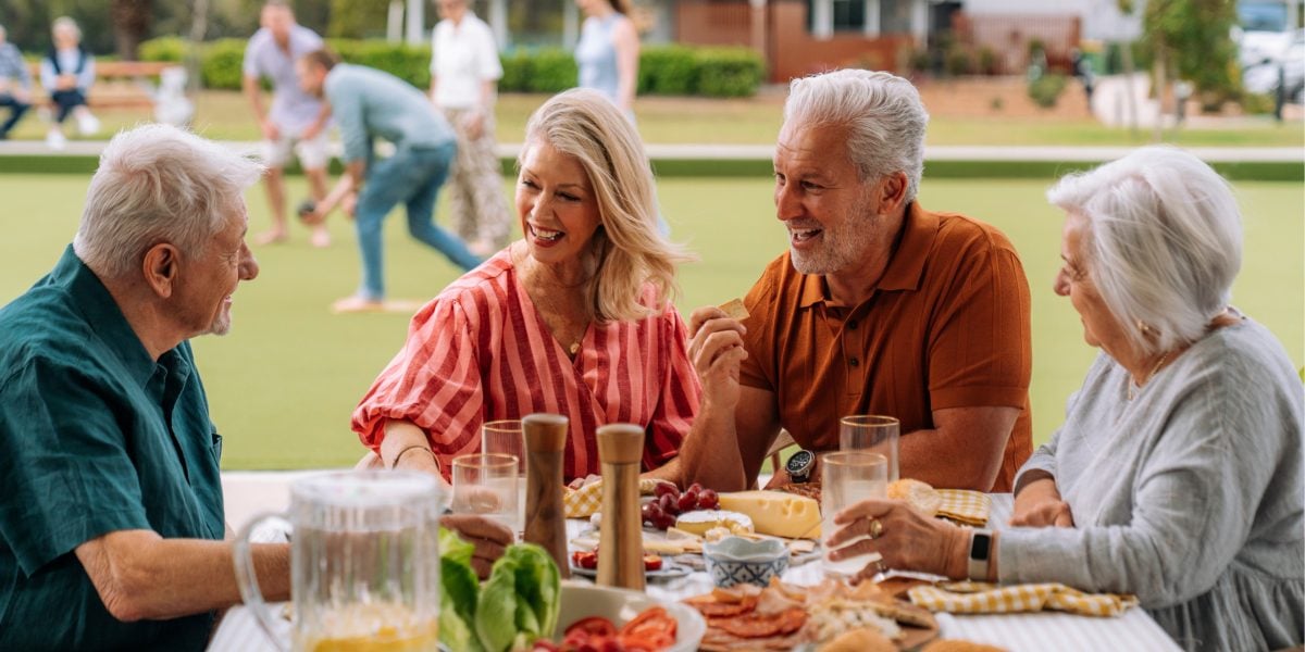 Four older adults sit at an outdoor table sharing food and laughing together, enjoying fresh vegetables and drinks. In the background, a park buzzes with people playing—a perfect day out for Over 50s Wangaratta.