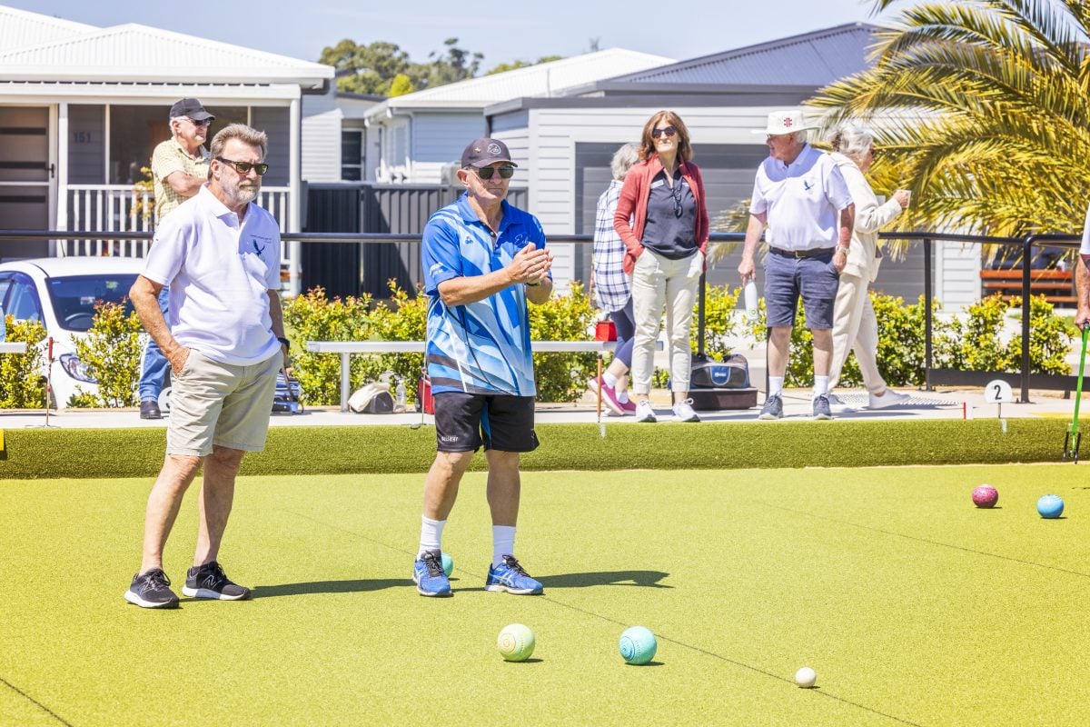 A group of people play lawn bowls on a sunny day, showcasing community partnerships. Two men stand on the green near several bowls, whilst others watch from the sidelines. Houses and greenery are in the background.