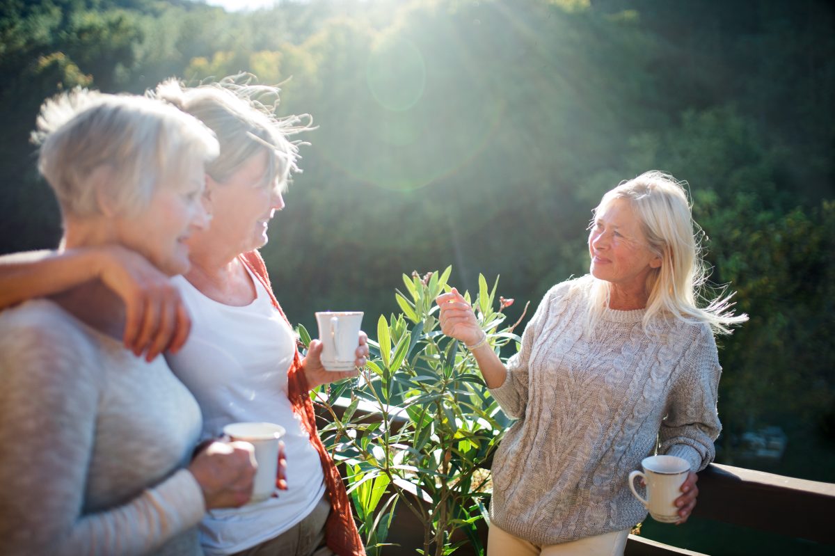 Three older women stand outside in sunlight, enjoying a relaxed lifestyle as they hold mugs and chat happily near green plants, with trees and nature in the background.