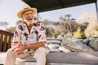 An older man wearing a straw hat and a colourful shirt with a van print sits outdoors on a cushioned bench, smiling and laughing. Trees and a mountain are visible in the sunny background.