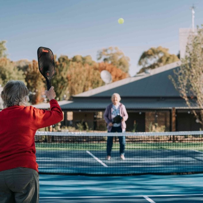 Two older adults embrace a vibrant lifestyle over 50s as they play pickleball on an outdoor court, one in a red jumper hitting the ball over the net while the other gets ready to return. Trees and a building form the backdrop.