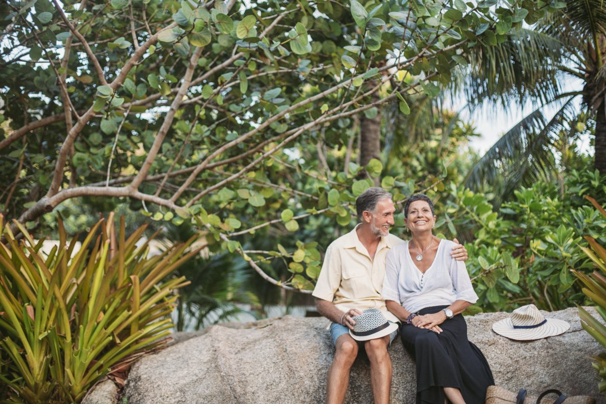 An older couple sits on a large rock surrounded by lush greenery, smiling and embracing. Both wear light, casual clothing and have hats beside them, enjoying a relaxed moment outdoors.