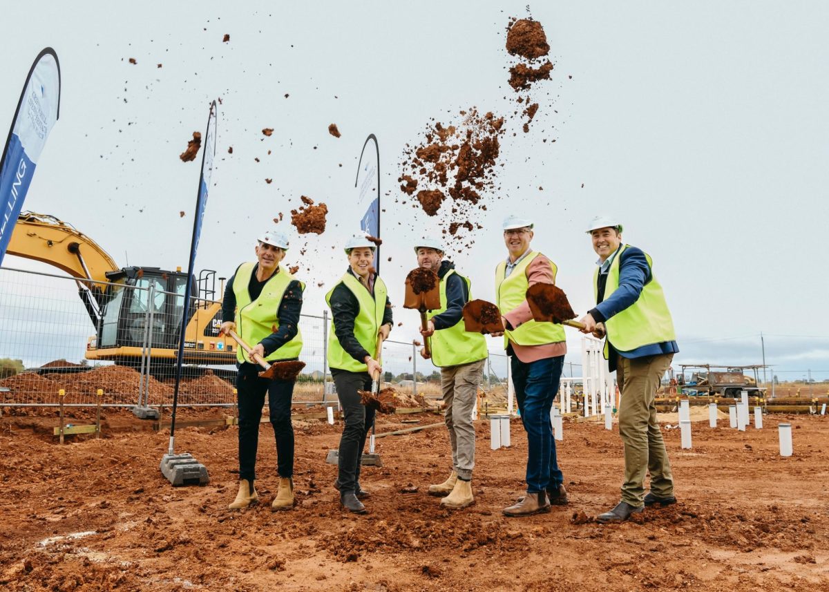 Five people in safety vests and hard hats use shovels to toss soil at a construction site, with an excavator and flags in the background. The ground is red earth, and white pipes stick up from the ground.