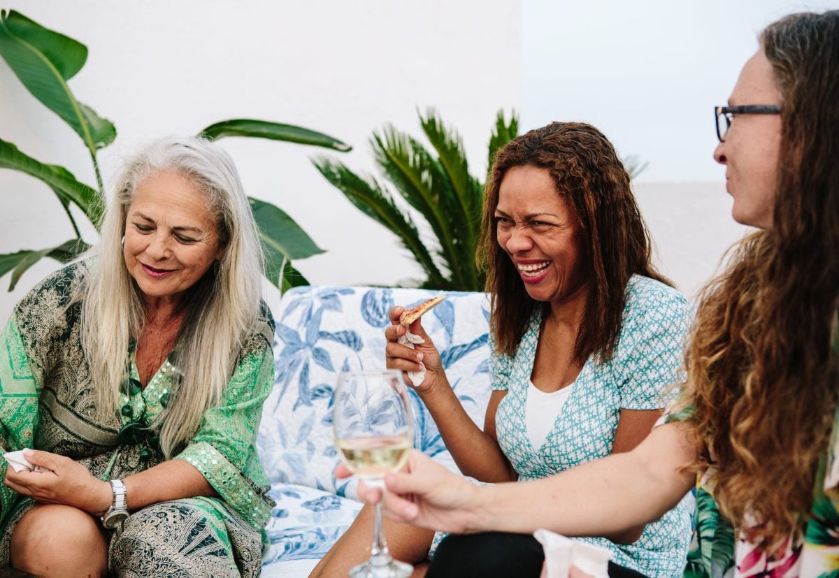 Three women sit together outdoors, laughing and enjoying food and drinks. One woman holds a slice of pizza, another has a glass of white wine. Surrounded by green plants and patterned cushions, they embody our difference through joy and connection.