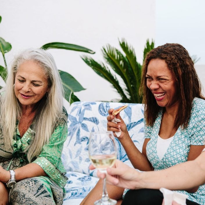 Three women sit together outdoors, laughing and enjoying food and drinks. One woman holds a slice of pizza, another has a glass of white wine. Surrounded by green plants and patterned cushions, they embody our difference through joy and connection.