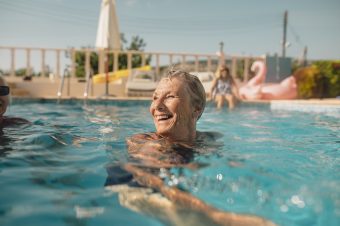 An older woman smiles while swimming in a pool on a sunny day, embracing a joyful lifestyle. In the background, a pink inflatable flamingo and another person sitting poolside add to the relaxed atmosphere.