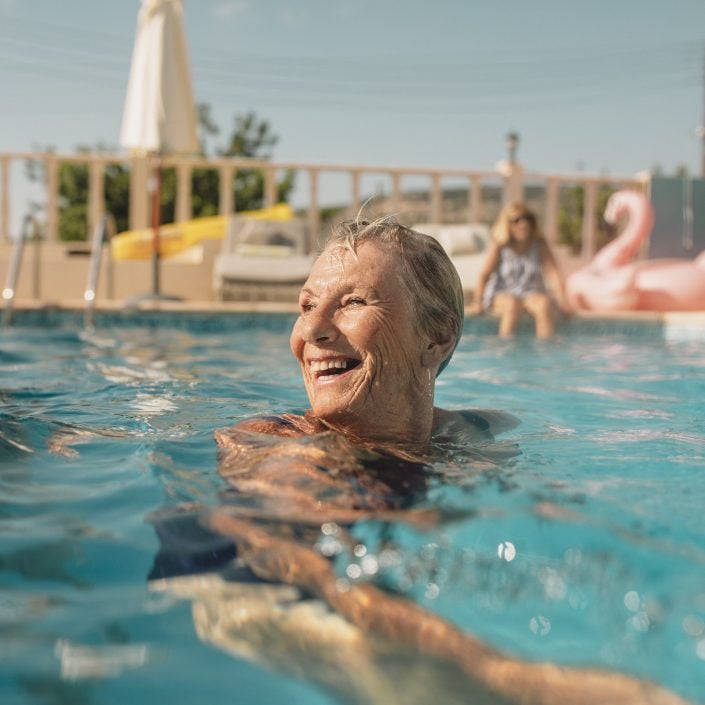 An older woman smiles while swimming in a pool on a sunny day, embracing a joyful lifestyle. In the background, a pink inflatable flamingo and another person sitting poolside add to the relaxed atmosphere.