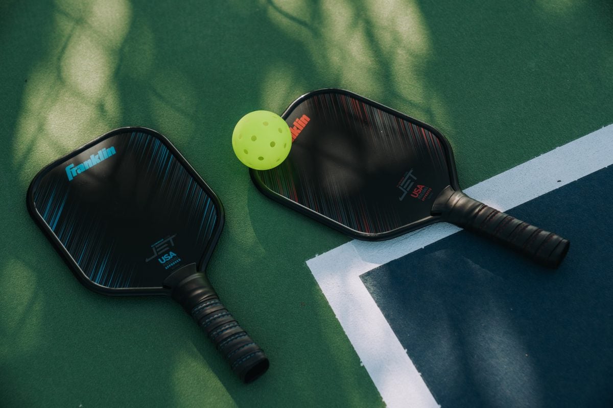 Two pickleball bats and a yellow perforated ball rest on a green court near a white boundary line, with dappled sunlight and shadows visible—perfect for enjoying the game while managing weekly site fees.