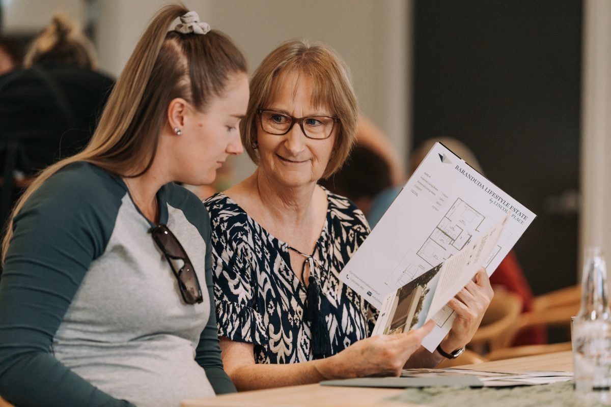 Two women sitting together, one showing the other some printed floor plans and documents. They appear to be discussing the papers in a casual indoor setting. One woman is smiling and wearing spectacles.