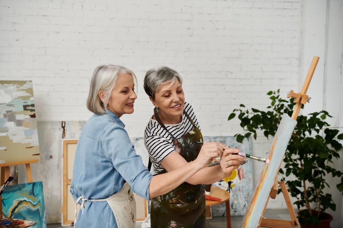 Two older women with grey hair happily painting together on a canvas in a bright art studio, wearing aprons. One woman guides the other's hand. Artwork and a large green plant are visible in the background.