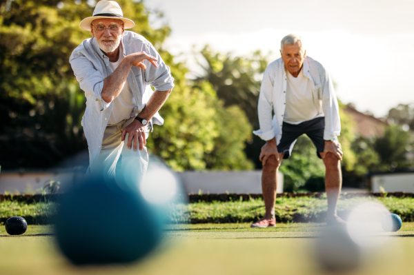Two older men playing lawn bowls outdoors on a sunny day; one is bowling a ball while the other watches, both dressed casually with trees and greenery in the background—enjoying leisure without worrying about weekly site fees.
