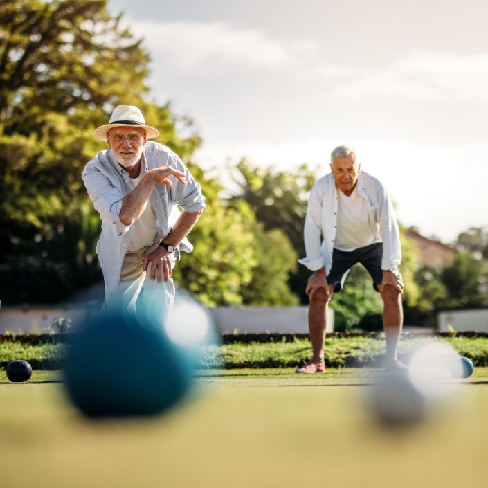 Two older men playing lawn bowls outdoors on a sunny day; one is bowling a ball while the other watches, both dressed casually with trees and greenery in the background—enjoying leisure without worrying about weekly site fees.