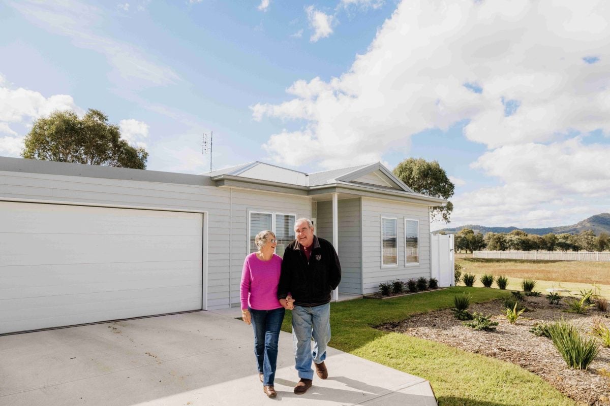 An older couple walk hand in hand, smiling, in front of a modern single-storey house with a garage and landscaped garden—embracing the vibrant lifestyle over 50s, set against mountains under a partly cloudy sky.