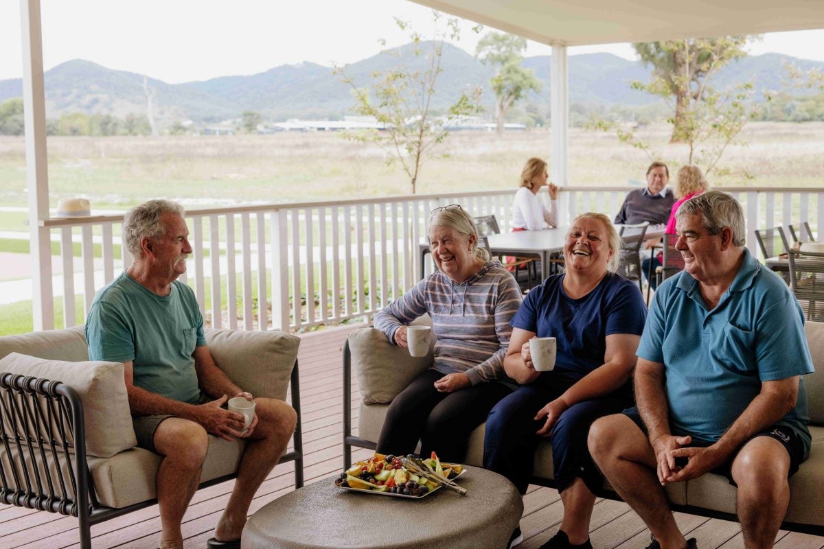 Four older adults sit together on a patio, smiling and holding mugs. They are relaxed and chatting, enjoying land lease living with mountains and fields visible in the background, and a plate of fruit on the table.