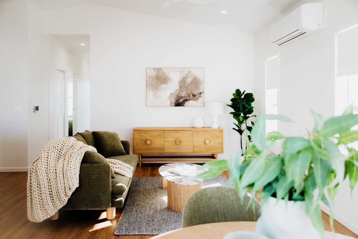 Bright, modern living room designed for a stylish lifestyle over 50s, with a green sofa, textured blanket, wooden sideboard, abstract wall art, large potted plant, round coffee table, and leafy plant against white walls and wooden floors.