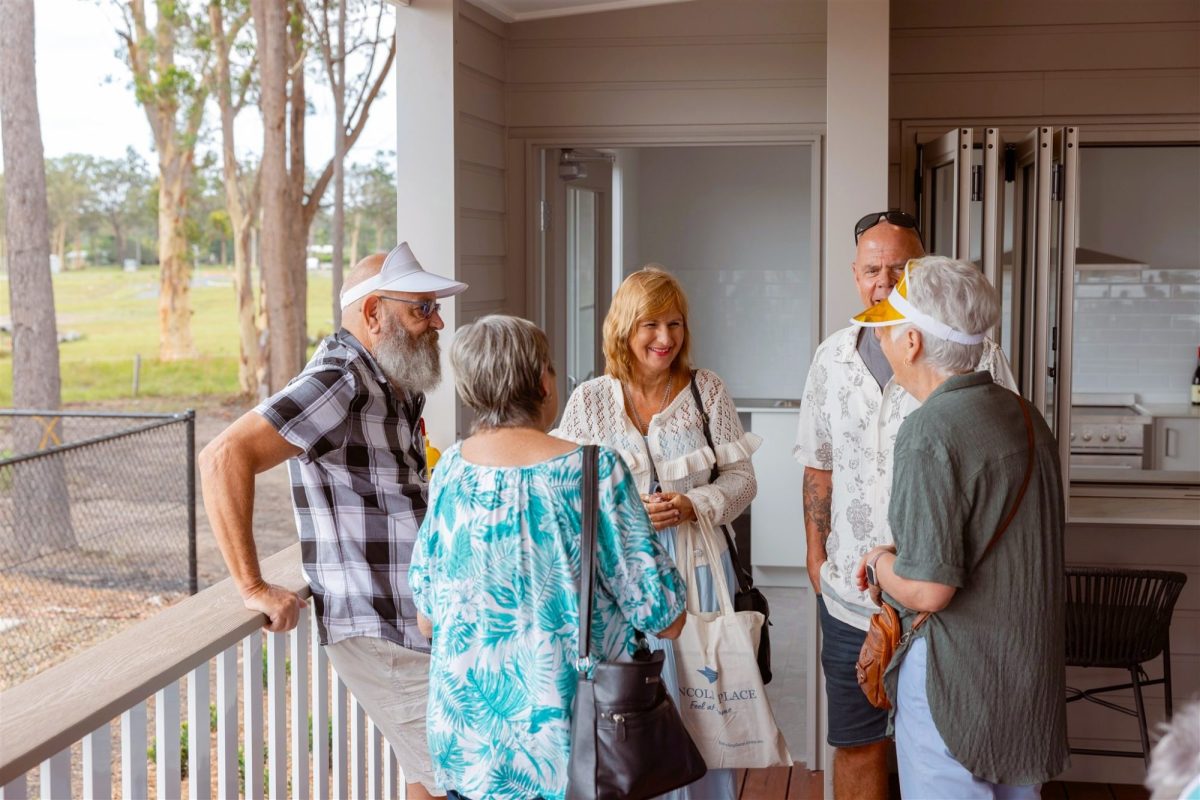 A group of five older adults stand and chat on a porch in a land lease living community. They are casually dressed, smiling, and appear to be enjoying each other’s company. Trees and a grassy area are visible in the background.