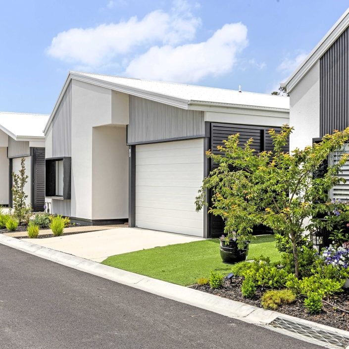 A row of modern, single-storey houses with white exteriors and gabled roofs lines a clean, paved street under a partly cloudy sky—an inviting community with well-maintained gardens and affordable weekly site fees.