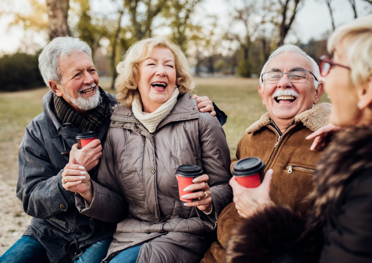 Four older adults sit on a bench outdoors at Campbell Lifestyle Estate, smiling and laughing together whilst holding takeaway coffee cups. They wear warm jackets and appear to be enjoying a cheerful conversation in the park.