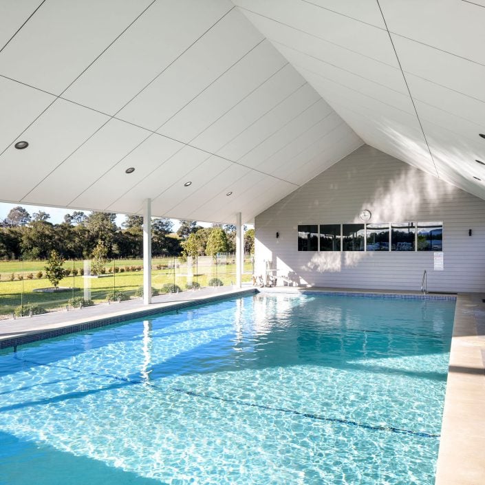 A large indoor swimming pool with clear blue water, modern white ceiling, and loungers along the side—perfect for relaxing after a day at the Campbell Bowls Tournament. Large windows overlook a grassy outdoor area with trees.