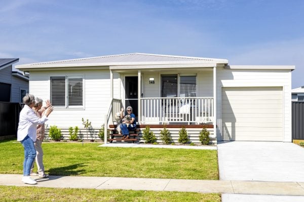 A modern single-storey house with a front porch. Two people sit on the steps with a dog, discussing the Campbell Bowls Tournament, while two others walk past on the pavement, waving. The garden is green and well-kept under a bright blue sky.