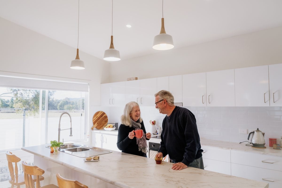 An older couple stands in a bright, modern kitchen, smiling and talking whilst holding mugs. Sunlight streams through a large window, illuminating the white cupboards and marble worktop.