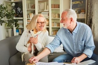 An elderly woman holding a small dog sits on a sofa next to an elderly man reviewing papers. Both are smiling and appear happy in a homely, book-filled living room after their Hunter Valley Smooth Moves session.