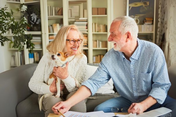 An elderly woman holding a small dog sits on a sofa next to an elderly man reviewing papers. Both are smiling and appear happy in a homely, book-filled living room after their Hunter Valley Smooth Moves session.