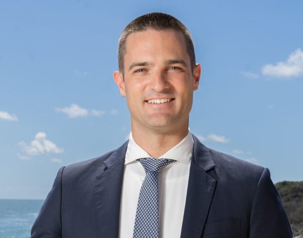 Daniel Kelly, dressed in a navy suit and patterned tie, smiles at the camera with a clear blue sky, some clouds, and the sea in the background.