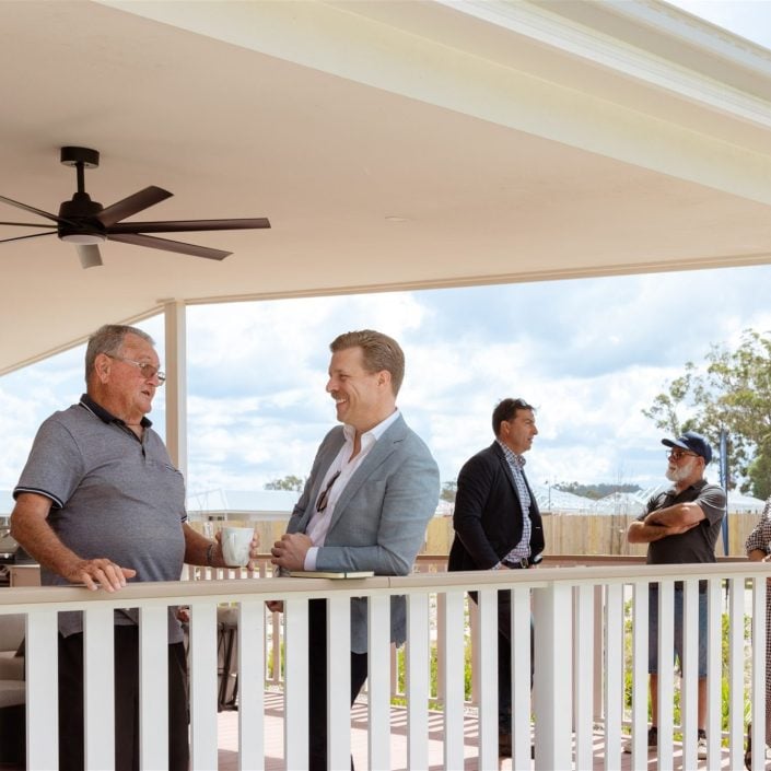 A group from the Northern Rivers over-50s Clubhouse stand and talk on a covered porch; three lean on the railing in conversation whilst others chat in the background, with trees and a cloudy sky behind them.